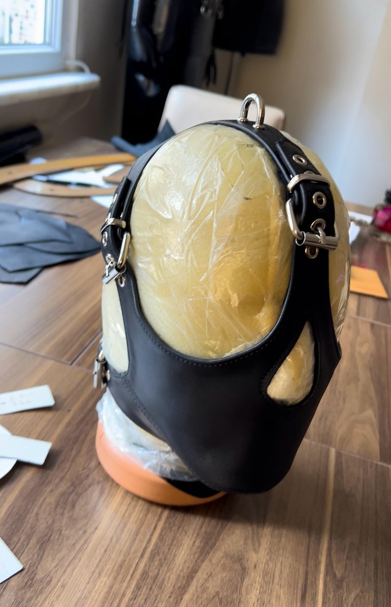 May include: Black leather mask with a cutout for the nose and mouth. The mask is attached to a black leather harness with silver buckles and rings.