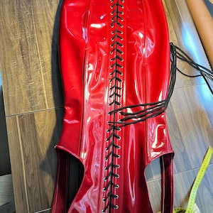 Könnte beinhalten: Ein glänzendes rotes Korsett mit schwarzer Schnürung in der Mitte. Das Korsett hat ein hoch tailliertes Design und besteht aus einem glänzenden Material. Das Kleidungsstück wird auf einer Holzoberfläche präsentiert.
