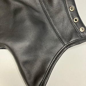 May include: Close-up of a black leather garment. The leather has a textured surface with visible stitching and metal eyelets along the edge. The garment appears to be a piece of clothing.