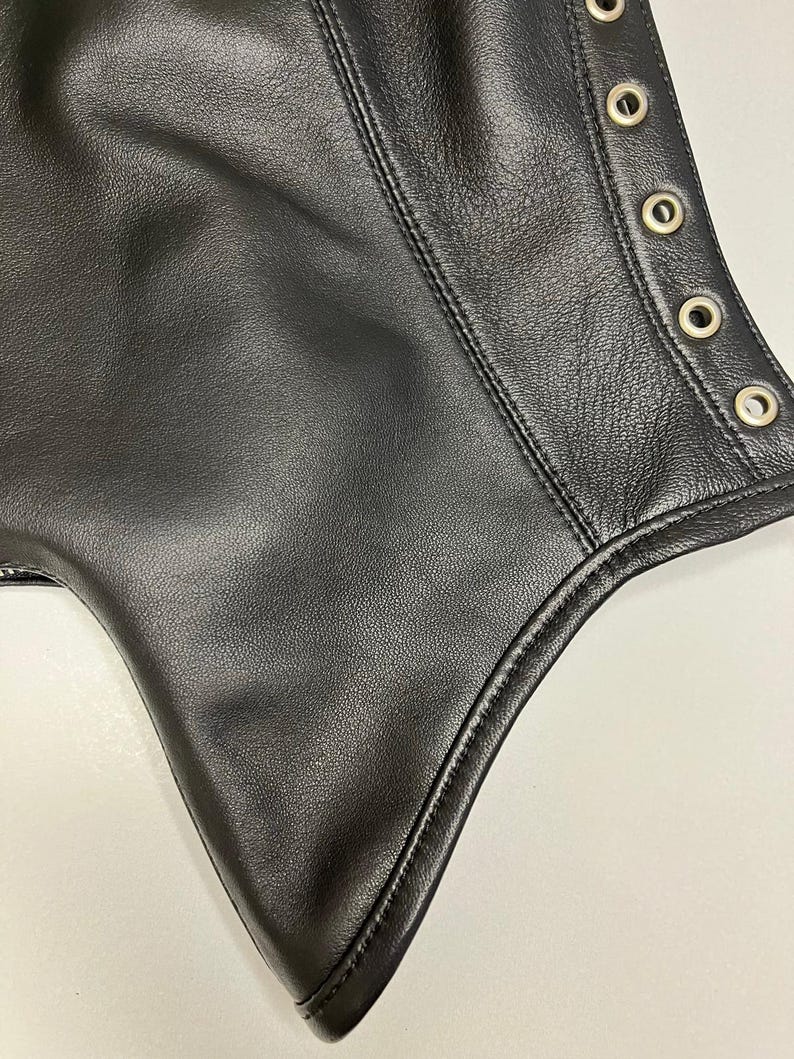 May include: Close-up of a black leather garment. The leather has a textured surface with visible stitching and metal eyelets along the edge. The garment appears to be a piece of clothing.
