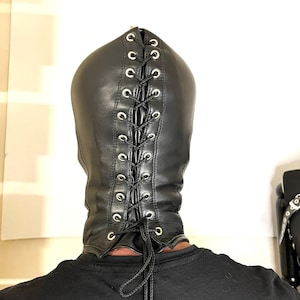 May include: Black leather hood with a lace-up back. The hood has silver-toned eyelets and a drawstring closure. The person is wearing a black shirt. The hood covers the head and neck.