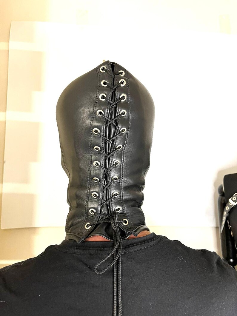 May include: Black leather hood with a lace-up back. The hood has silver-toned eyelets and a drawstring closure. The person is wearing a black shirt. The hood covers the head and neck.