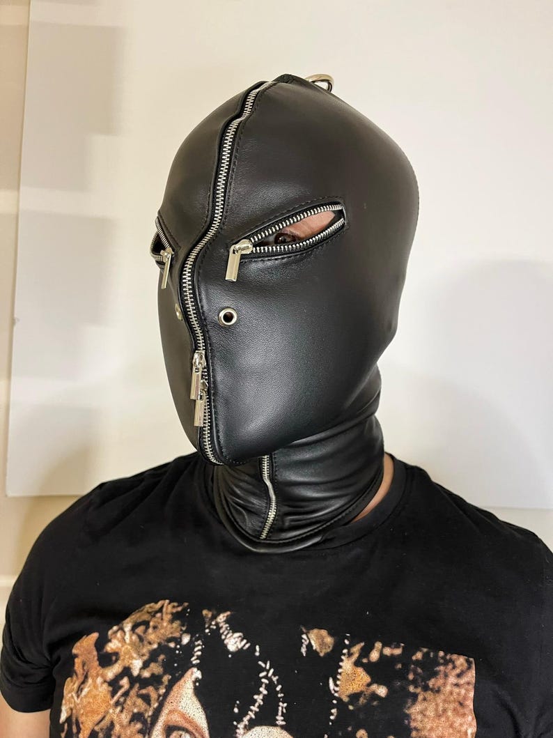May include: Black leather hood with multiple silver zippers. The hood covers the entire head and neck, with openings for the eyes. The zippers run vertically and horizontally, adding a unique design element to the hood.
