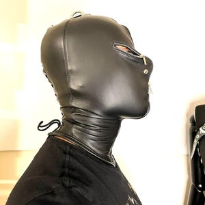May include: Black leather hood with a zipper across the mouth and a metal ring on top. The hood covers the head and neck, with a laced closure at the back. The design features a smooth, sleek texture.
