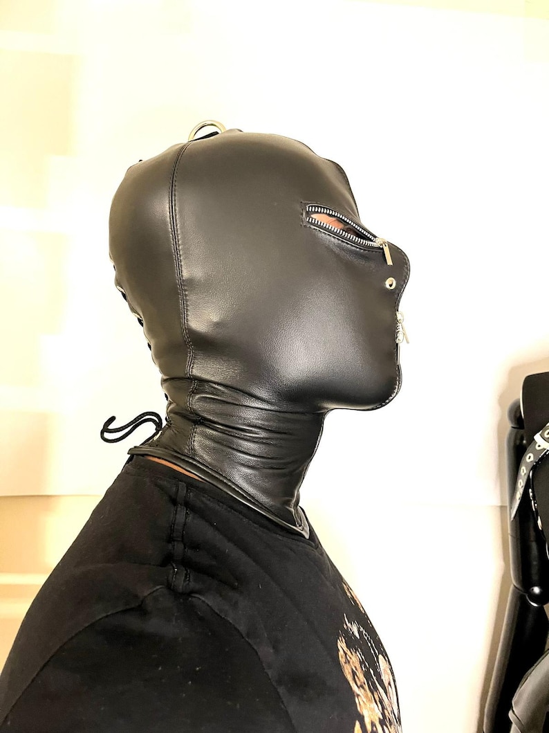 May include: Black leather hood with a zipper across the mouth and a metal ring on top. The hood covers the head and neck, with a laced closure at the back. The design features a smooth, sleek texture.