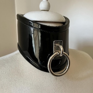 Może przedstawiać: Czarny choker z lakierowanej skóry z przymocowanym srebrnym pierścieniem. Choker znajduje się na białej głowie manekina.