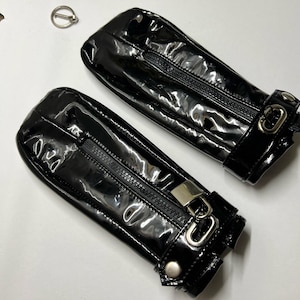 Könnte beinhalten: Paar schwarze, glänzende, fingerlose Handschuhe mit Reißverschlüssen und silberfarbener Hardware. Die Handschuhe haben einen Schnallenverschluss und ein kleiner Metallring ist ebenfalls vorhanden.