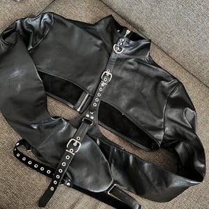May include: Black leather jacket with a cropped bodice and long sleeves. The jacket has a zip fastening, a high collar, and multiple straps with silver buckles and eyelets.