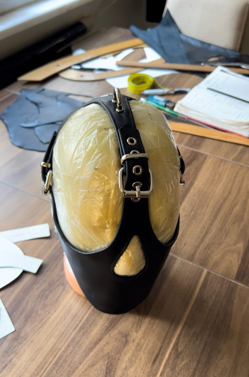 May include: Black leather mask with a strap and buckles. The mask is on a mannequin head.