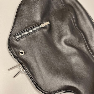 May include: Black leather bag with a curved shape. Features a silver zipper closure and two silver metal accents. The bag has a smooth texture and a minimalist design.