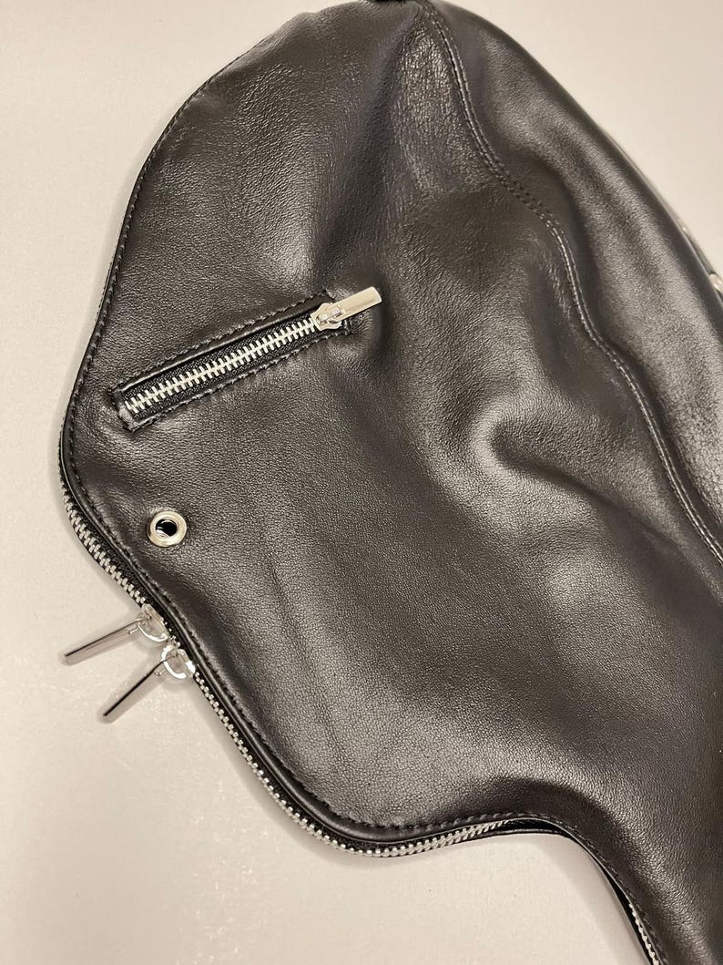 May include: Black leather bag with a curved shape. Features a silver zipper closure and two silver metal accents. The bag has a smooth texture and a minimalist design.
