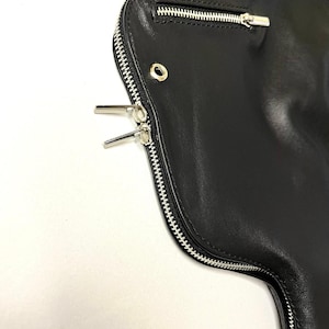 May include: Close-up of a black leather jacket with silver zippers. The jacket features a curved design with a main zipper and a smaller pocket zipper. The leather appears smooth and the zippers are detailed.