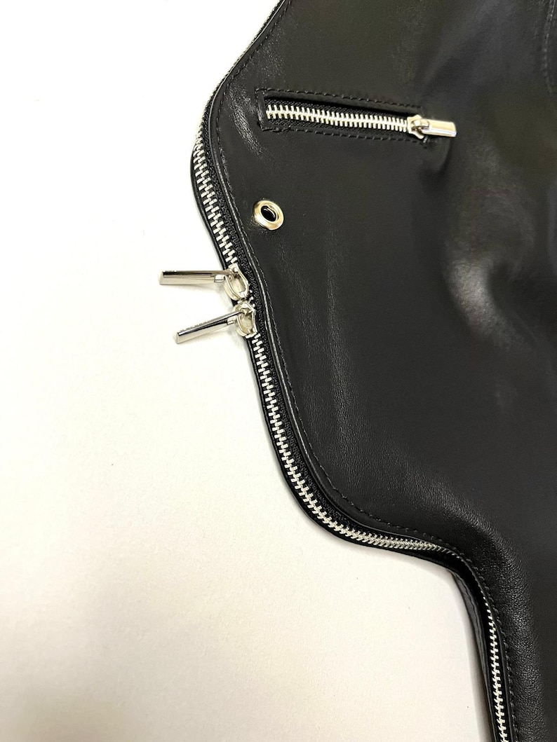 May include: Close-up of a black leather jacket with silver zippers. The jacket features a curved design with a main zipper and a smaller pocket zipper. The leather appears smooth and the zippers are detailed.