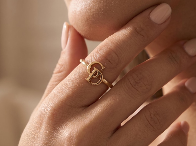 K&ouml;nnte beinhalten: Ein goldfarbener Ring mit einem stilisierten, ineinandergreifenden Monogramm-Design mit den Buchstaben S und D. Der Ring hat ein d&uuml;nnes, poliertes Band und wird an einem Finger getragen, was das metallische Finish und die kursive Schrift zeigt.