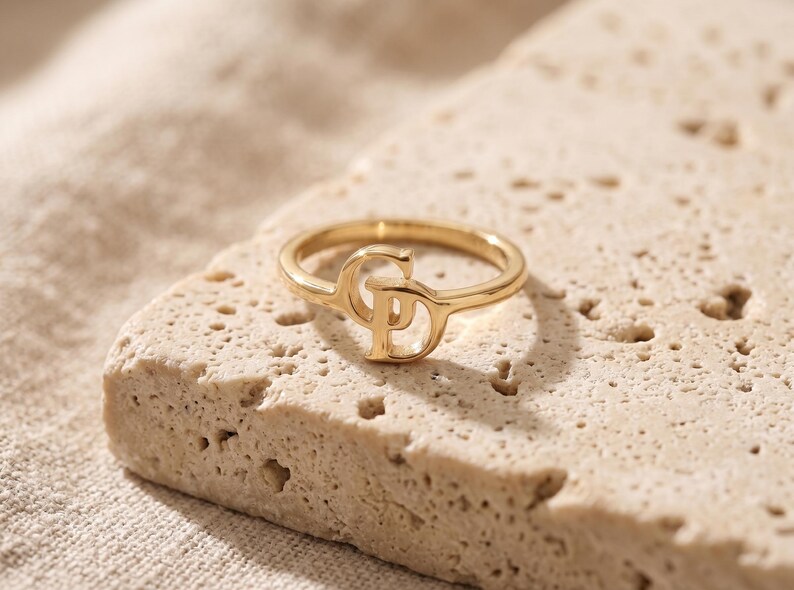 K&ouml;nnte beinhalten: Ein goldfarbener Ring mit einem zentralen Design aus den ineinandergreifenden Gro&szlig;buchstaben C und P. Der Ring ist d&uuml;nn und kreisf&ouml;rmig, wobei die Buchstaben in den oberen Teil des Rings integriert sind und eine durchgehende Metallform bilden.
