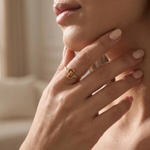 K&ouml;nnte beinhalten: Ein goldfarbener Ring mit einem stilisierten Buchstaben D als zentralem Blickfang. Die Ringschiene ist d&uuml;nn und kreisf&ouml;rmig und aus einem polierten Metall gefertigt, das das Licht reflektiert. Das Design ist schlicht und besteht aus dem Buchstaben.