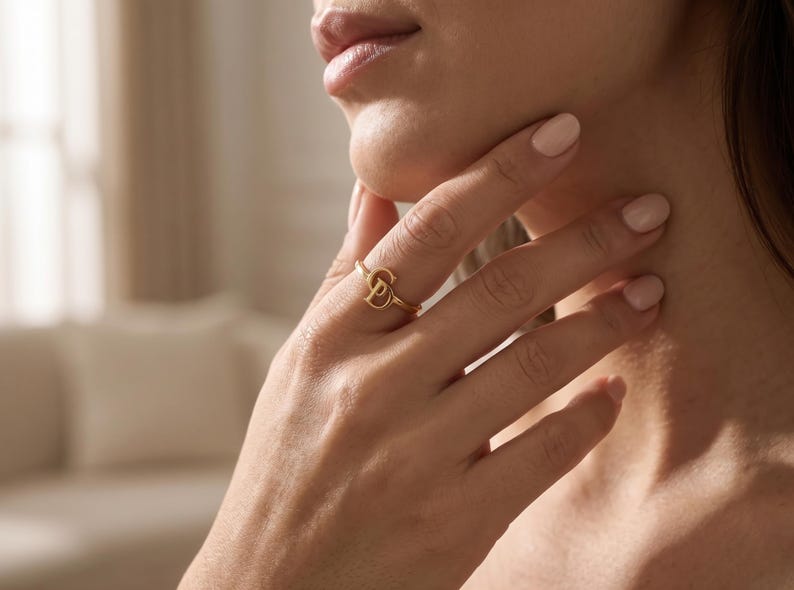 K&ouml;nnte beinhalten: Ein goldfarbener Ring mit einem stilisierten Buchstaben D als zentralem Blickfang. Die Ringschiene ist d&uuml;nn und kreisf&ouml;rmig und aus einem polierten Metall gefertigt, das das Licht reflektiert. Das Design ist schlicht und besteht aus dem Buchstaben.