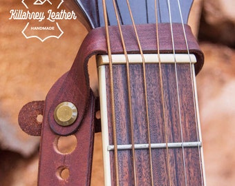 May include: A close-up of a brown leather guitar strap with a brass buckle. The strap is attached to a guitar with a wooden neck and metal strings. The strap has the text "Killaney Leather Handmade" on it.