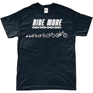 Può includere: Una maglietta blu scuro con il testo "RIDE MORE" in lettere bianche. Sotto il testo è stampata una catena di pignoni di bicicletta, con una progressione di biciclette da un triciclo a una mountain bike.