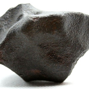 Pode incluir: Um meteorito de cor cinza escuro, de forma irregular, com uma superfície áspera e texturizada.
