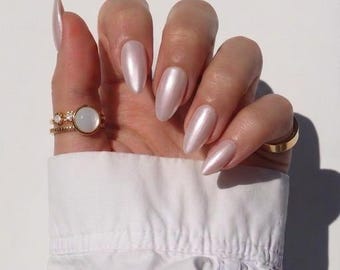 Puede incluir: Un conjunto de dos anillos de oro con una piedra blanca redonda central y piedras de acento más pequeñas, junto a una banda de oro lisa en el dedo anular. Las uñas tienen forma de almendra puntiaguda y un acabado de esmalte nacarado.