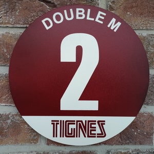 May include: A circular, burgundy metal sign featuring the text "DOUBLE M" above a large white number "2". The word "TIGNES" is displayed in a white band at the bottom. The sign is mounted on a brick wall, likely indicating a property number.