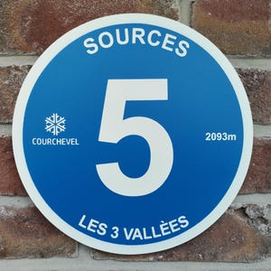 May include: A circular blue sign with a white border featuring the large white number 5 in the centre. The text reads SOURCES at the top, LES 3 VALLEES at the bottom, and COURCHEVEL with a snowflake icon and 2093m on the sides.