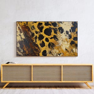 Puede incluir: Una consola de medios de madera con tres compartimentos que presentan un frente de rat&aacute;n tejido. La consola tiene un acabado de madera clara y se asienta sobre patas inclinadas. Una pintura abstracta enmarcada con un esquema de color amarillo, marr&oacute;n y negro cuelga sobre la consola.