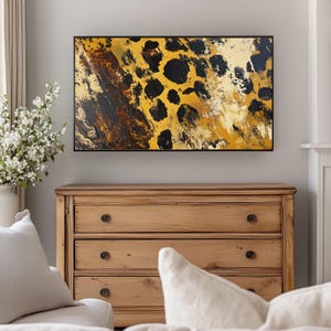 Puede incluir: Pintura abstracta de un estampado de leopardo en tonos de amarillo, marr&oacute;n y negro. La pintura est&aacute; enmarcada en un marco negro y est&aacute; colgada en una pared sobre una c&oacute;moda de madera.
