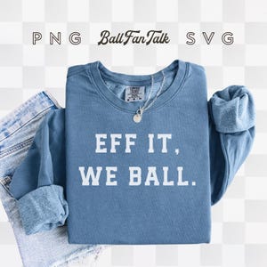 以下が含まれることがあります： 「EFF IT, WE BALL」の文字が入った青いスウェットシャツ。スウェットシャツは袖を折りたたんであります。ペンダント付きのシルバーのネックレスがスウェットシャツの上にあります。青いジーンズが一部見えています。
