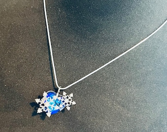 Op de afbeelding: Een zilveren ketting met een blauwe kristallen sneeuwvlok hanger. De sneeuwvlok is gemaakt van kleinere kristallen en heeft een hartvorm in het midden.