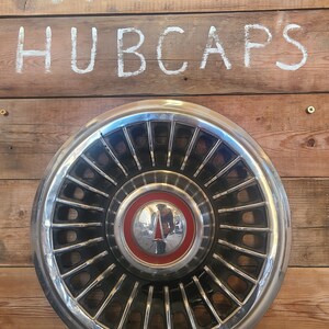May include: A vintage chrome hubcap with a black centre and a red and chrome emblem. The hubcap has a spoke design and is mounted on a wooden surface with the words "HELM HUBCAPS" painted in white.