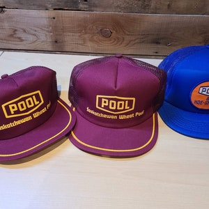 May include: Three trucker hats with mesh back panels. Two are burgundy with yellow "POOL Saskatchewan Wheat Pool" text and one is blue with yellow "POOL & HOE-GRASS" text.