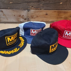 Op de afbeelding: Vier baseballpetten met het logo "MF Massey Ferguson" geborduurd op de voorkant. De petten zijn in verschillende kleuren: blauw, zwart, rood en zwart met een gouden bladontwerp.