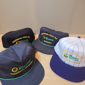 May include: Four blue and white baseball caps with the Saskatchewan Wheat Pool logo and Westco branding. The caps have a green and yellow logo with the text "Saskatchewan Wheat Pool" and "Westco" embroidered on the front.