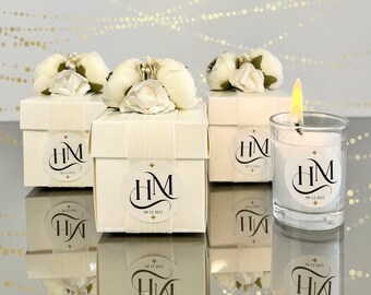 May include: Four white favor boxes with white flowers on top and a white sticker with the initials "HM" and the date "08 12 2022". A clear glass candle with a white sticker with the initials "HM" and the date "08 12 2022".