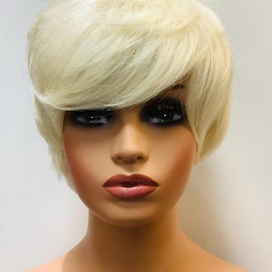May include: A mannequin head with a short, layered blonde wig. The wig has a side-swept bang and is styled to frame the face. The mannequin has full makeup, including dark eye shadow and pink lipstick. The skin tone is light.