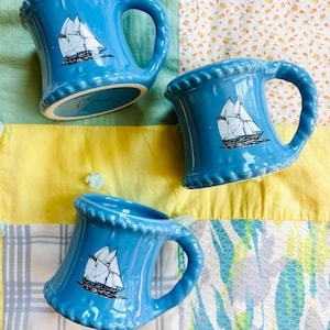 May include: Three bright blue ceramic mugs with a flared base and a rope-textured rim. Each mug features a black and white graphic illustration of a sailing ship with multiple masts and white sails centred on the front of the glossy blue surface.