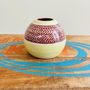 May include: A small, round ceramic vase with a textured, reddish-brown pattern on the upper half. The vase has a yellow base and a yellow band near the top, with a dark brown interior. The vase is displayed on a wooden surface.