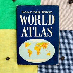 May include: A vintage World Atlas book with a blue cover and the title "WORLD ATLAS" in large white letters. The cover features a map of the world in orange and yellow. The book is on a patchwork background of green, yellow, tan, and blue gingham fabric.