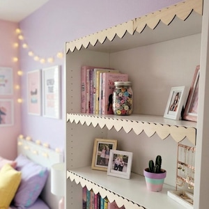May include: White bookshelf with scalloped edges, filled with books, framed photos, and a jar of colorful buttons. A small potted cactus sits on a shelf. The room has a soft, pastel color scheme with fairy lights.