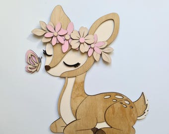 May include: Wooden deer wall art featuring a deer with a flower crown and a butterfly. The deer is made of light brown wood with pink and beige accents. The butterfly is pink and beige. The deer is sitting down.