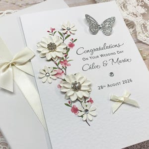 May include: A white wedding card with the text "Congratulations On Your Wedding Day Chloe & Martin 28th August 2026". It features white paper flowers, a silver butterfly, and a small bow. The card is in a white box with a cream ribbon.
