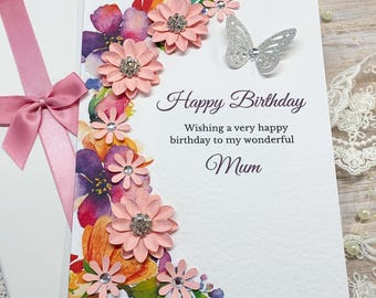 Może przedstawiać: Biała kartka urodzinowa z kwiatowym wzorem i napisami "Happy Birthday" i "Mum". Karta jest ozdobiona różowymi, fioletowymi i pomarańczowymi kwiatami oraz srebrnym motylem. Karta jest w białym pudełku z różową wstążką.