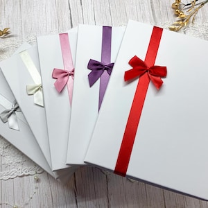 May include: Five white gift boxes, each adorned with a satin ribbon and bow in various colours: silver, cream, pink, purple, and red. The ribbons are tied diagonally, offering a decorative and elegant appearance.