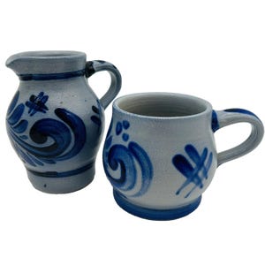 May include: A light gray ceramic pitcher and a matching mug, both featuring hand-painted dark blue swirling floral patterns and cross-hatch designs. The pitcher has a curved handle and a pouring spout, while the mug has a rounded handle.
