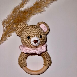 May include: A crocheted brown bear rattle with pink accents. The bear has a wooden ring attached to its body.