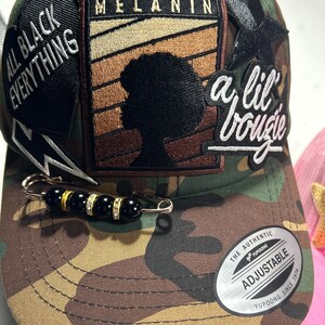 May include: Camouflage baseball cap with black and white patches. The patches say "All Black Everything", "Melanin", and "a lil bougie". The cap is adjustable and has a black and gold safety pin with black beads.