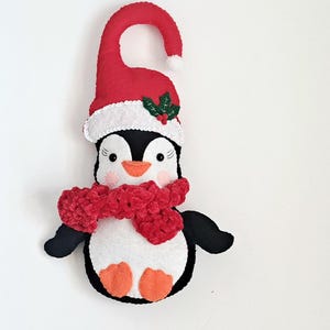 May include: A felt penguin door hanger featuring a red Santa hat with a white pom-pom and green holly leaves. The penguin has a black and white body, an orange beak and feet, and a textured red crocheted scarf wrapped around its neck.