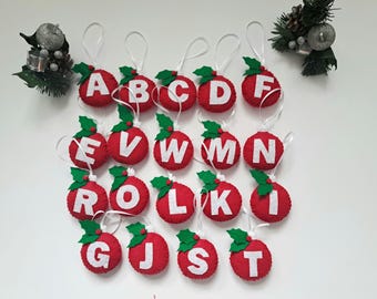 May include: Red felt Christmas ornaments, each with a white letter from the alphabet. Green holly leaves and white ribbon hang from the top. The ornaments are arranged in rows. Decorative holiday accents are in the background.